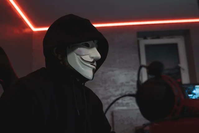 Person wearing a hacker mask in a dimly lit room, symbolizing cyber security. Impersonation risks: where the loss lands, and for whom